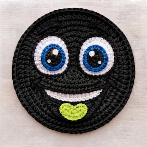 May include: A circular black crochet coaster featuring a cartoon face with large white and blue eyes, a wide white smile, and a small lime green heart-shaped tongue protruding from the mouth, all crafted with detailed yarn stitching.