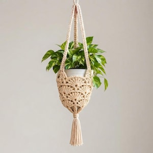 Pode incluir: Um suporte de plantas em crochê bege com um padrão de leques e uma borla longa na parte inferior. O suporte contém um vaso branco com uma planta pothos de folhas verdes em forma de coração, suspenso por uma alça de corda trançada no topo.