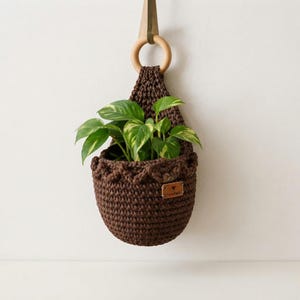 May include: A dark brown crocheted hanging planter with a scalloped top edge and a light wooden ring at the top for suspension. A small rectangular leather tag with a heart icon and text is attached to the front, containing a green pothos plant.