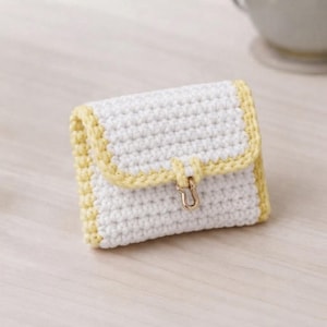 May include: A small, rectangular crochet pouch featuring a white body with a textured stitch pattern. The edges are trimmed in a soft yellow yarn. A gold-toned metal clasp is centered on the front flap, securing the closure of the accessory.