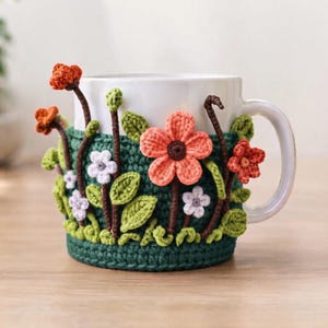 May include: A white ceramic mug featuring a dark green crocheted cozy. The cozy is decorated with three-dimensional crocheted flowers in shades of orange, coral, and white, along with green stems and leaves attached to the exterior surface.