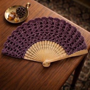 Puede incluir: Un abanico con estructura de madera clara con detalles calados y una parte superior de encaje de ganchillo en color morado oscuro. El encaje presenta un borde festoneado y un delicado patrón floral calado en toda la tela.