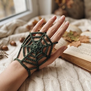 Könnte beinhalten: Ein dunkelwaldgrünes Häkel-Handaccessoire mit einem komplizierten Spinnennetzmuster. Das Stück ist so gestaltet, dass es den Handrücken bedeckt, mit einer Schlaufe, die um den Mittelfinger führt, und einem Band, das das Handgelenk umschließt.