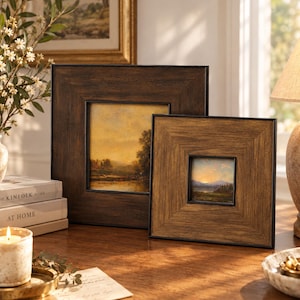 May include: Two square picture frames with dark brown wood grain textures. The larger frame features a landscape painting of a river at sunset, while the smaller frame displays a mountain landscape at dusk. Both frames have wide, beveled borders.