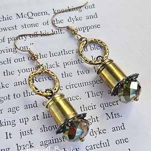 May include: A pair of steampunk-style earrings. Each earring features a brass bullet casing, a faceted iridescent crystal bead, and a textured gold-tone ring. The earrings hang from gold-colored ear wires. The background is a page of printed text.