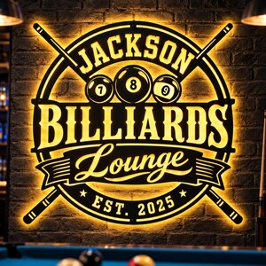 May include: A circular black metal wall sign with warm yellow LED backlighting. It features the text JACKSON BILLIARDS Lounge EST. 2025, crossed pool cues, and three billiard balls numbered 7, 8, and 9 centered within the design.