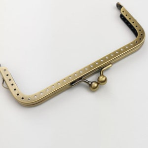 Puede incluir: Un marco de bolso de metal dorado con dos cierres redondos. El marco está diseñado para ser utilizado en la elaboración de bolsos y carteras.