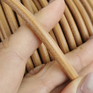 Puede incluir: Primer plano de un carrete de cordón de cuero marrón claro. El cordón está enrollado firmemente alrededor del carrete y está siendo sujetado por una mano.