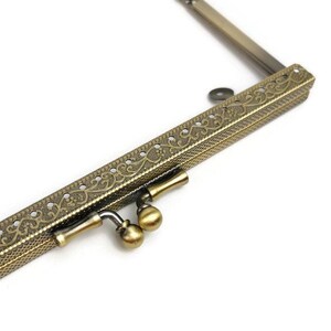 May include: A gold-toned metal purse frame with an ornate design. The frame has two clasps and a decorative edge.