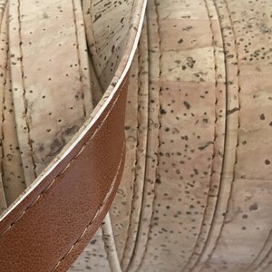 May include: Close-up of a brown leather strap with white stitching, folded over a beige cork material with stitching. The cork has a natural, textured surface.