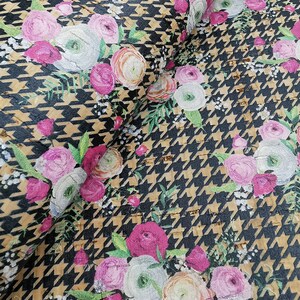May include: Fabric with a black and tan houndstooth pattern, overlaid with clusters of pink, white, and peach-colored floral bouquets. Green leaves and small white flowers accent the floral arrangements. The fabric is folded, showing its texture.