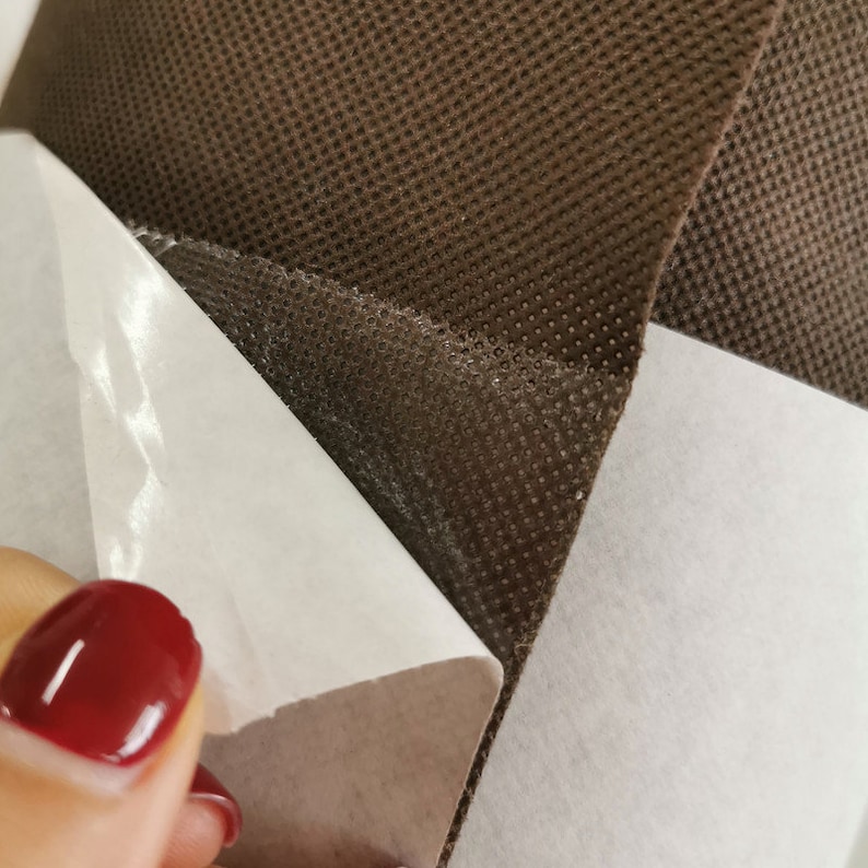 May include: A close-up of brown fabric with a white adhesive backing. The fabric has a textured, dotted pattern. The adhesive backing is partially peeled back to reveal the sticky surface.