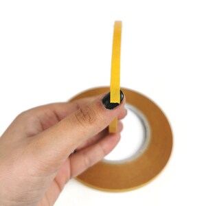 Puede incluir: Un rollo de cinta amarilla con respaldo marr&oacute;n. La cinta est&aacute; siendo sostenida por una mano.