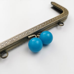 Puede incluir: Primer plano de un marco de bolso de metal con un diseño decorativo. El marco es de color bronce antiguo y tiene dos cuentas de color azul turquesa unidas a la parte inferior.