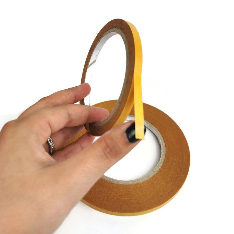 Puede incluir: Un rollo de cinta adhesiva de doble cara con un lado adhesivo amarillo y un respaldo de papel marr&oacute;n.