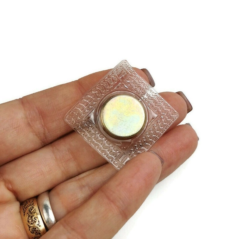 May include: A clear plastic square with a round, silver-colored magnet in the center. The magnet is surrounded by a decorative pattern.