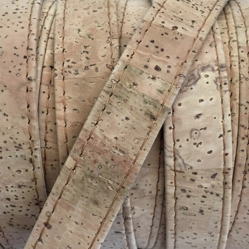 May include: Close-up of a roll of natural cork ribbon with a stitched edge. The ribbon is a light brown color with a speckled pattern.