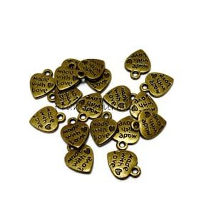 Puede incluir: Un grupo de 18 dijes de metal de color dorado en forma de corazón con las palabras "Made with Love" y "Love Apow" impresas en ellos. Los dijes tienen un bucle en la parte superior para sujetarlos a joyas u otras manualidades.