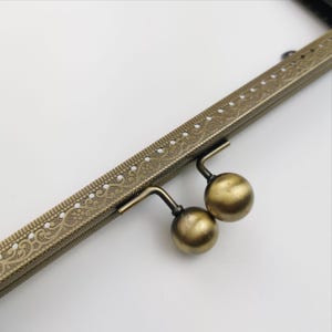 Op de afbeelding: Een close-up van een gouden metalen tasframe met een sierlijk ontwerp. Het frame heeft twee ronde, decoratieve knoppen.