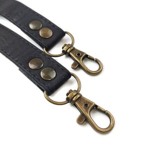 May include: Close-up of two black leather straps with antique brass-colored swivel clasps and rivets. The straps are connected to the clasps with round metal rings. The background is white.