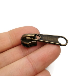 Op de afbeelding: Antiek gouden rits-trekker met een lus aan het einde. De rits-trekker is gemaakt van metaal en heeft een licht verweerde afwerking.