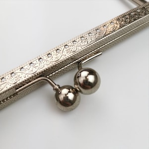 Può includere: Primo piano di una cornice per borsa in metallo argentato con un design decorato e due chiusure rotonde in metallo.