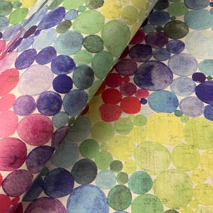 May include: A roll of patterned fabric with a repeating design of watercolour-style circles. The circles are in shades of red, pink, purple, blue, green, and yellow, creating a rainbow effect. The fabric has a textured appearance.