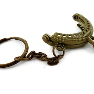 Puede incluir: Un marco de bolso de color bronce con un llavero adjunto. El marco está diseñado para ser utilizado para un bolso pequeño o una cartera.