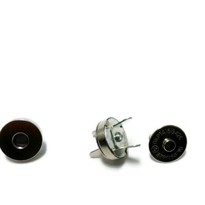 Puede incluir: Tres cierres magnéticos plateados para proyectos de costura. Un cierre es un disco redondo con un pequeño agujero en el centro. Los otros dos cierres son un par de cierres magnéticos con tres puntas cada uno. Los cierres tienen el texto "OKPTA 516426" impreso en ellos.