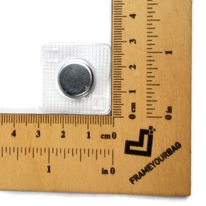 May include: A close-up of a small, clear, square-shaped magnetic button with a silver-colored magnet in the center. The button is measured against a ruler, showing it is approximately 1 centimeter in size.