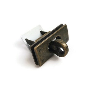 Puede incluir: Cierre de metal de tono latón antiguo con un marco rectangular y un anillo para sujetar una correa o cadena. El cierre tiene un acabado texturizado y un diseño decorativo.