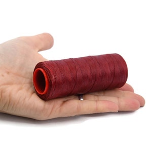May include: A spool of red waxed thread for sewing or leather crafting. The thread is wound around a plastic spool with a red centre.