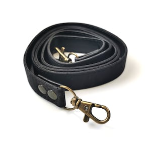 May include: A coiled black strap with antique brass-colored metal clasps and rivets. The strap is made of a textured material and is designed for use with a bag or purse.