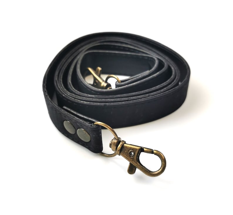 May include: A coiled black strap with antique brass-colored metal clasps and rivets. The strap is made of a textured material and is designed for use with a bag or purse.