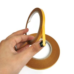 Puede incluir: Un rollo de cinta adhesiva de doble cara con un lado adhesivo amarillo y un respaldo de papel marr&oacute;n.