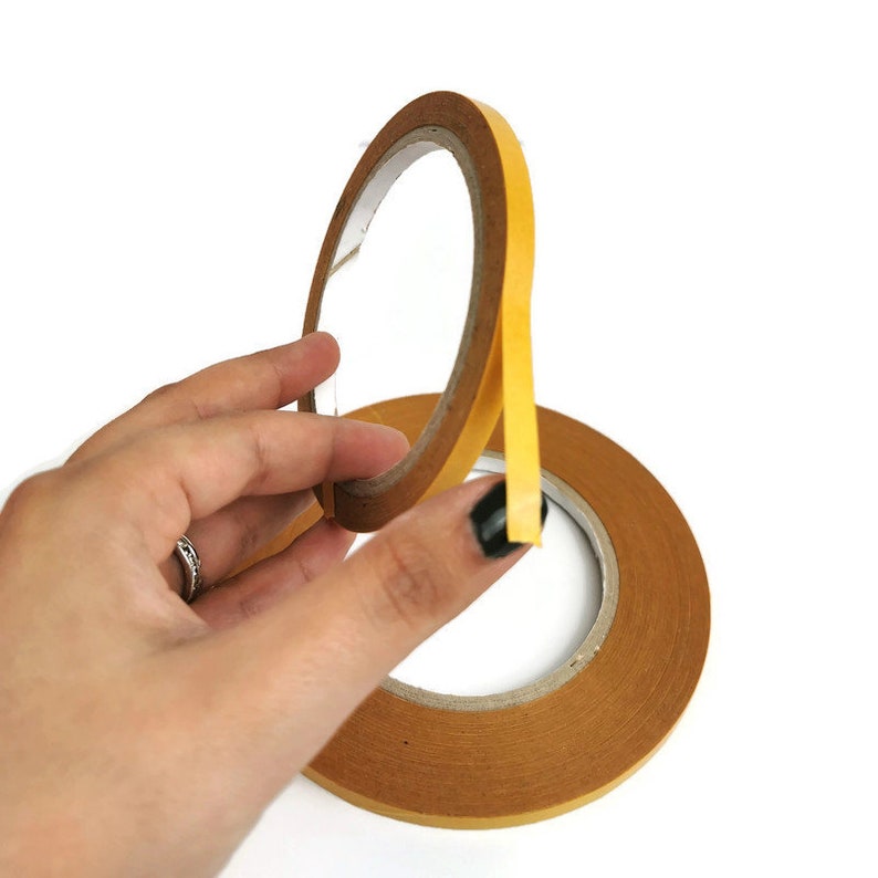 Puede incluir: Un rollo de cinta adhesiva de doble cara con un lado adhesivo amarillo y un respaldo de papel marr&oacute;n.