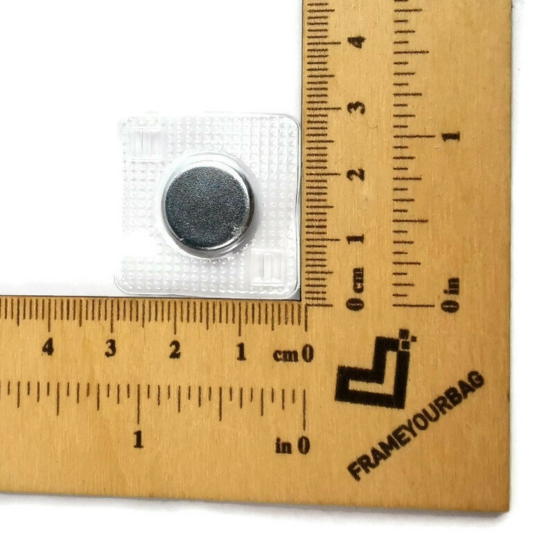 May include: A close-up of a clear plastic magnetic snap closure with a silver metal disc. The snap is measured against a ruler, showing it is approximately 1 cm in size.