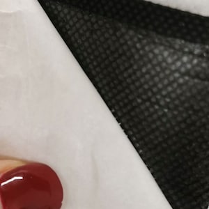 Peut inclure: Gros plan d'un masque facial noir avec une surface textur&eacute;e, recouvrant partiellement une surface blanche. Une petite partie d'un ongle peint en rouge est visible dans le coin inf&eacute;rieur gauche.