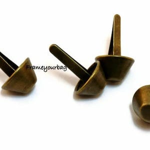 Pode incluir: Rebites de metal com cor de latão antigo com base em forma de cone e poste retangular. Os rebites são projetados para fixação em tecido ou couro. A imagem mostra vários rebites, alguns com o poste estendido e outros com o poste dobrado.