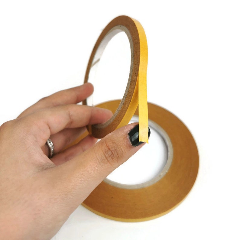 Puede incluir: Un rollo de cinta adhesiva de doble cara con un respaldo amarillo y un adhesivo transparente. La cinta se est&aacute; sujetando con una mano, y el extremo de la cinta se est&aacute; despegando.