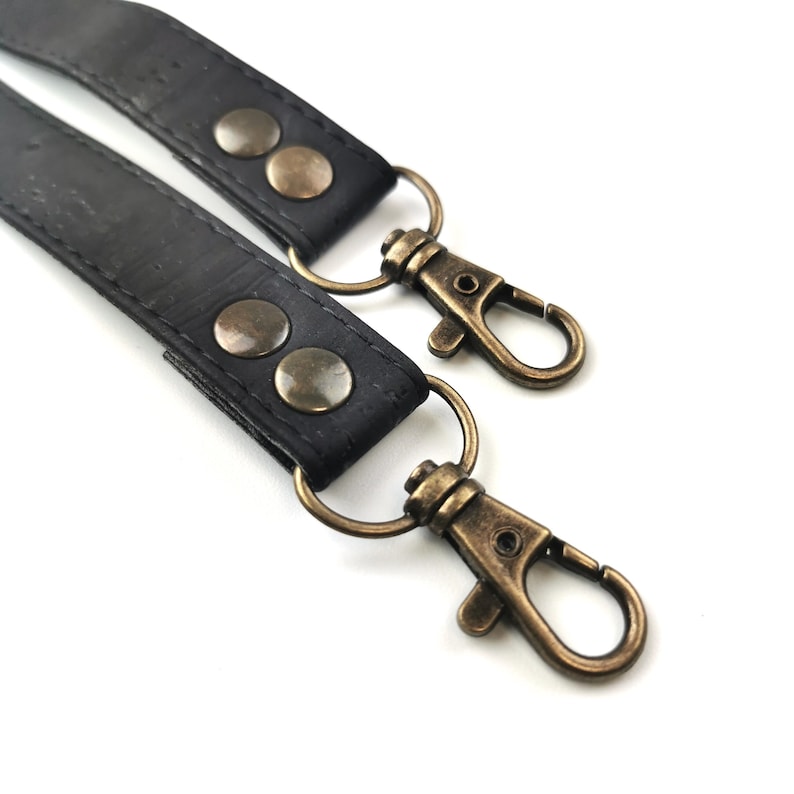 May include: Close-up of two black leather straps with antique brass-colored swivel clasps and rivets. The straps are connected to metal rings. The background is white.