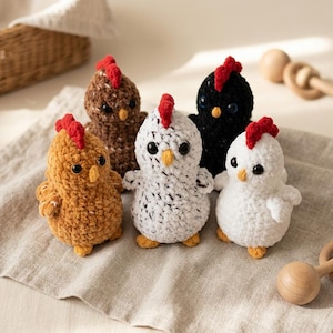 Op de afbeelding: Vijf kleine gehaakte kippenknuffels in diverse kleuren, waaronder bruin, wit met zwarte spikkels, effen wit en zwart. Elk knuffeltje is voorzien van een felrode kam, een klein geel snaveltje en ronde zwarte oogjes, gemaakt van zacht garen.