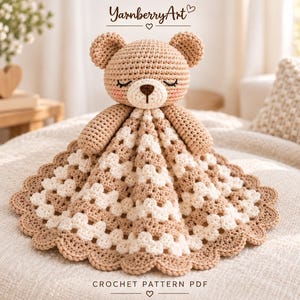 Peut inclure: Une couverture de sécurité au crochet avec une tête d'ours beige aux yeux fermés, un nez marron et des joues roses. La couverture attachée présente un motif de carrés granny texturé en rayures alternées crème et beige avec une bordure festonnée.