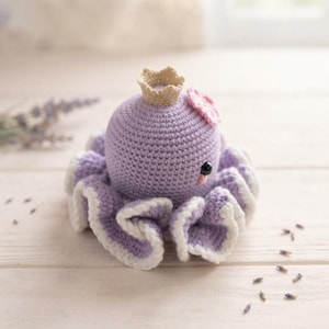 May include: A crocheted octopus toy in a light purple color with a white ruffled edge along the base. The octopus features a small beige crown on top, a pink flower accent on the side, and two black bead eyes with a small pink stitched mouth.