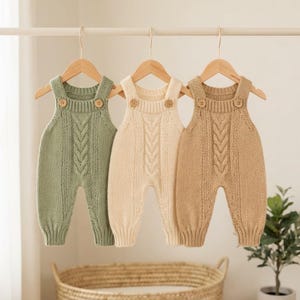 May include: Three knitted overalls hanging on wooden hangers. The overalls are sage green, cream, and tan. Each piece features a central vertical cable knit pattern, ribbed leg cuffs, and two small wooden buttons on the front shoulder straps.