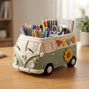 May include: A crochet desk organizer shaped like a vintage van in sage green and cream. The van features a yellow sunflower appliqué on the side, a string of colorful triangular bunting, and black wheels. It is filled with various colored pencils and pens.