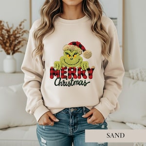 May include: A sand-colored crewneck sweatshirt featuring a graphic of a green character wearing a red and black plaid Santa hat. The character is positioned above the red and black plaid text "MERRY" and the green cursive text "Christmas".