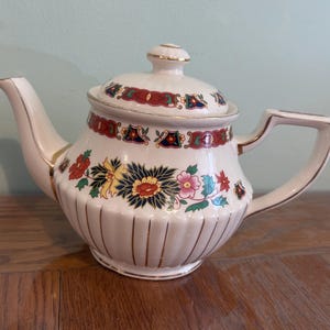 Può includere: Una teiera in ceramica color crema con base scanalata e linee verticali dorate. Il corpo e il coperchio sono decorati con un motivo floreale colorato in rosso, giallo, rosa e blu scuro, rifiniti con bordi dorati su manico, beccuccio e pomello.