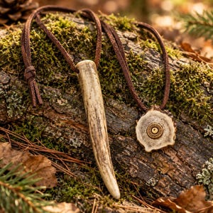 Può includere: Una collana con due pendenti su un cordino di cuoio marrone. Un pendente è una punta di corno lunga, liscia e di colore chiaro. Il secondo pendente è una fetta circolare di corno con un bossolo di metallo color ottone al centro.