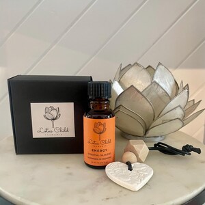 May include: A still life arrangement featuring an essential oil blend, a decorative lotus flower, and a heart-shaped ornament. The oil bottle has an orange label with the text "Energy". The heart ornament is white with a floral design.